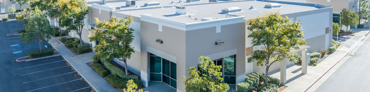 an angle-down view of a tan building with white trim and accents. Some trees and bushes make up the landscaping around the building with some parking area showing in the bottom left corner